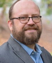 Dr. Feodor Gostjev smiling with short balding reddish brown hair, mustache and beard, wearing black rim glasses and a gray blazer over a light blue button up shirt