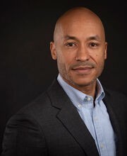 headshot of Dr. Jakari Griffith smiling with no hair and black and gray stubble mustache and beard wearing a blue and white pinstripe button down shirt under a dark gray suit jacket
