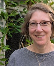 Dr. Laura K. Gross in the BSU Greenhouse smiling with short light brown hair and wearing brown rim glasses and a gray boat neck top.