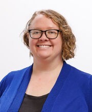 Ashley Hansen-Brown smiling with short wavy light brown hair and wearing black rim glasses and a royal blue sweater over a black blouse