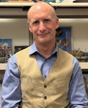 Loren Herren leaning on his desk smiling and wearing jeans, a blue button down shirt and beige vest