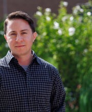 Dr. Asa Iacobucci standing outside in front of greenery with short brown hair wearing a black and purple check button up shirt