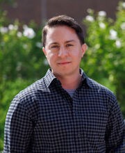 Dr. Asa Iacobucci standing outside in front of greenery with short brown hair wearing a black and purple check button up shirt