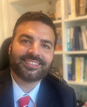 Dr. Chris H. Kennedy sitting in a desk chair with a built in book shelf in background. He is smiling, has short dark brown hair, beard and mustache and is wearing a navy blue suit jacket over a light blue button down shirt and red, white and blue tie