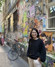 Inkyoung Kim smiling and standing next to a colorful building and a bicycle. She has medium length straight dark hair and is wearing white pants and black long sleeved top