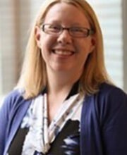 Dr. Heather Marella smiling with medium length blonde hair wearing brown rim glasses and a vneck black white and purple print dress with a purple jacket