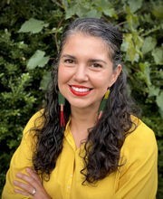 Dr. Alicia Mendez smiling in front of greenery with long graying dark brown hair pulled back on top and wearing a dark yellow button up blouse and long colorful earrings