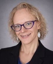 Dr. Brooke Merritt smiling with medium length curly blonde hair and wearing purple rim glasses, a blue blouse and black blazer