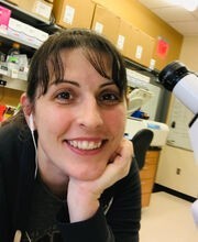 Dr. Joslyn Mills next to microscope in lab smiling with long dark brown hair pulled back in a pony tail with bangs and wearing earphones and a black long sleeve top