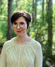 Dr. Sarah Fawn Montgomery standing in front of a wooded background with short dark brown hair and wearing round black rim glasses and a pale yellow cable knit sweater