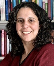Dr. Rachel Navarre smiling in front of a bookcase with long curly dark brown hair and wearing a v-neck maroon sweater