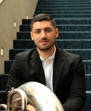 Jordan Oliveira sitting on a carpeted stairway with his tuba. He has short dark brown hair, mustache and beard and is wearing a black suit over a white button down shirt
