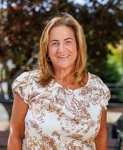 Kathleen Perry standing in front of a bench on campus smiling with medium length brown hair with blonde highlights and wearing an off-white with brown floral print short sleeve top