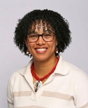 Cleonie Mainvielle smiling with medium length curly black hair and brown rim glasses and wearing a white and beige v-neck sweater