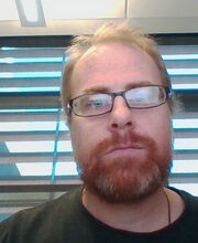 Dr. John F. Santore with reddish blonde receding hair, mustache and beard wearing brown rim glasses and a dark green shirt