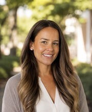 Dr. Hannah Taverna smiling with long brown wavy hair and wearing a gray open sweater over a white v-neck blouse