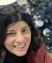 Sarah Thomas smiling with shoulder length wavy dark brown hair wearing a pair of purple rimmed glasses on top of her head and a red blazer over a black top