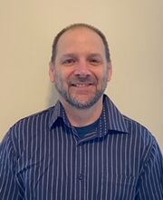 Dr. Stephen Waratuke smiling with balding brown hair and graying beard and mustache. He is wearing a navy button down shirt with white pinstripes over a navy t-shirt