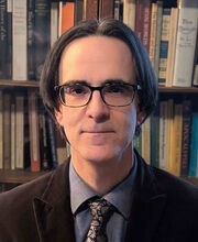 Dr. J.R. Webb with medium length salt and pepper straight hair wearing brown rim glasses, a blue button down shirt with a dark brown blazer and dark brown paisley tie