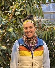 Emma Welch standing in a greenhouse smiling with brown hair in a messy bun on top of her head wearing a gold headband, gray long sleeve sweatshirt over an LLBean puffy vest with blue collar and orange, gold and beige stripes. She also wears 2 fashion scarves in colors of purple, gold and orange