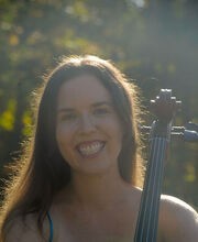 Dr. Sara Wilkins smiling with sun shining on her long dark brown hair. She is wearing a blue spaghetti strap top and the neck of her cello is in front of her shoulder