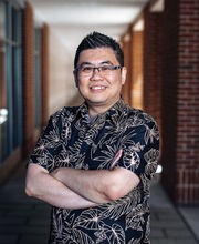 Dr. Cory Yeo smiling with short spiked black and gray hair wearing black rim glasses and a black short sleeve button down shirt with tropical leaves outlined in beige