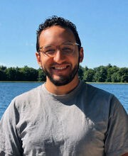 Dr. Alex Yepez smiling with short dark curly hair and a short dark beard and mustache. He is wearing light rimmed glasses and a gray short sleeve shirt and standing in front of a body of water with trees in background.