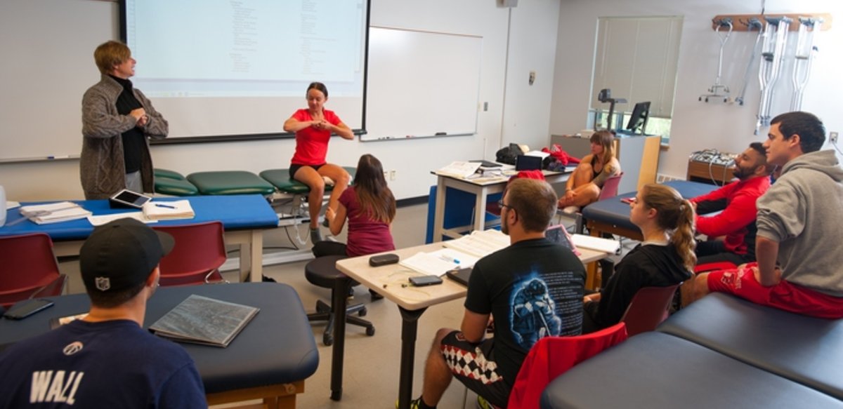 A class with desks and padded tables. At the front of the class a student sits on a padded table with her hands clasped together. The professor is standing next to the table with her hands similarly clasped. Another student is holding the first student's ankle. A class with desks and padded tables. At the front of the class a student sits on a padded table with her hands clasped together. The professor is standing next to the table with her hands similarly clasped. Another student is holding the first student's ankle.
