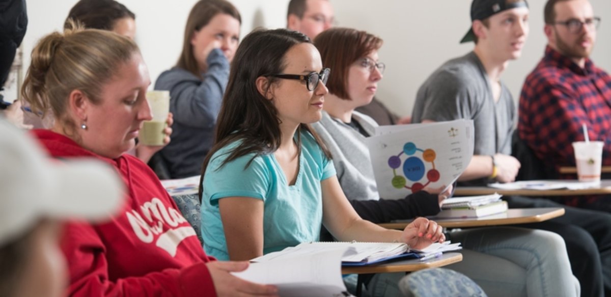 Students sitting in 2 rows of desks with notebooks out listening to a professor. Students sitting in 2 rows of desks with notebooks out listening to a professor.
