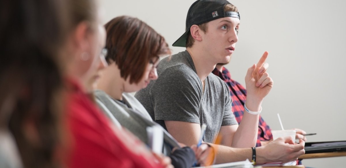 A student sitting in a row of desks with other students is pointing and speaking. A student sitting in a row of desks with other students is pointing and speaking.