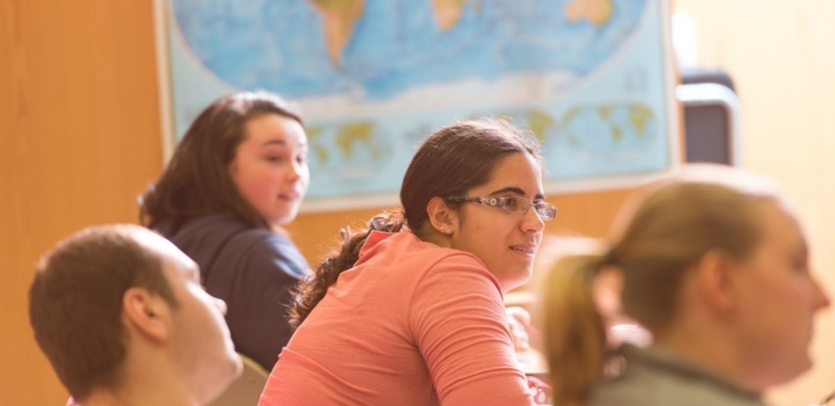 Four students sitting at a long table with a wall map in the background listening to a professor Four students sitting at a long table with a wall map in the background listening to a professor