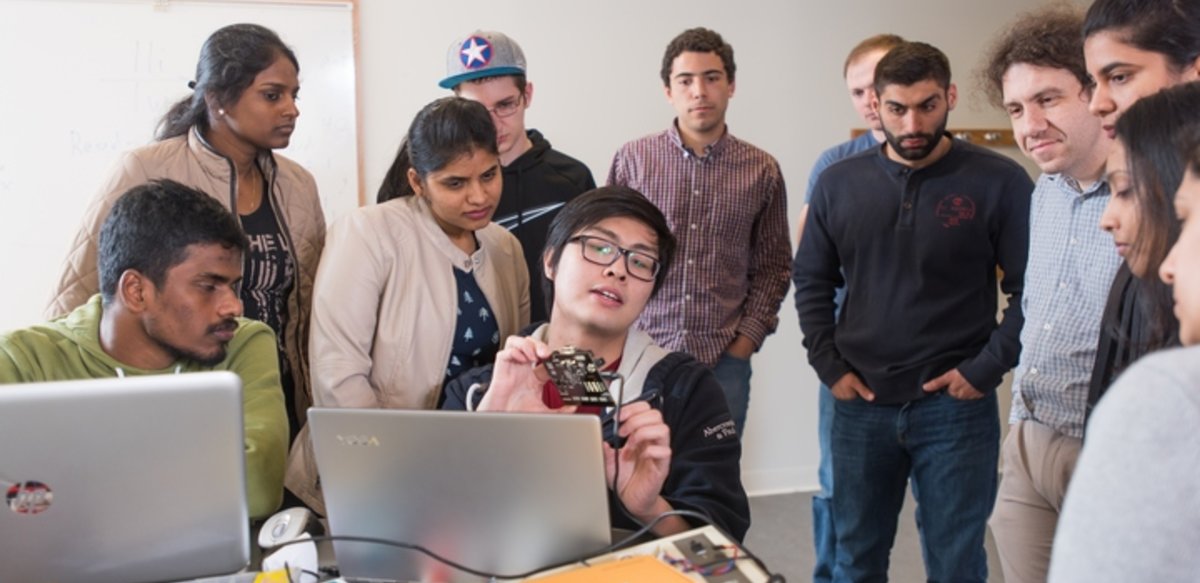 A student sitting at a desk showing his work to the professor and other students who are surrounding him. A student sitting at a desk showing his work to the professor and other students who are surrounding him.