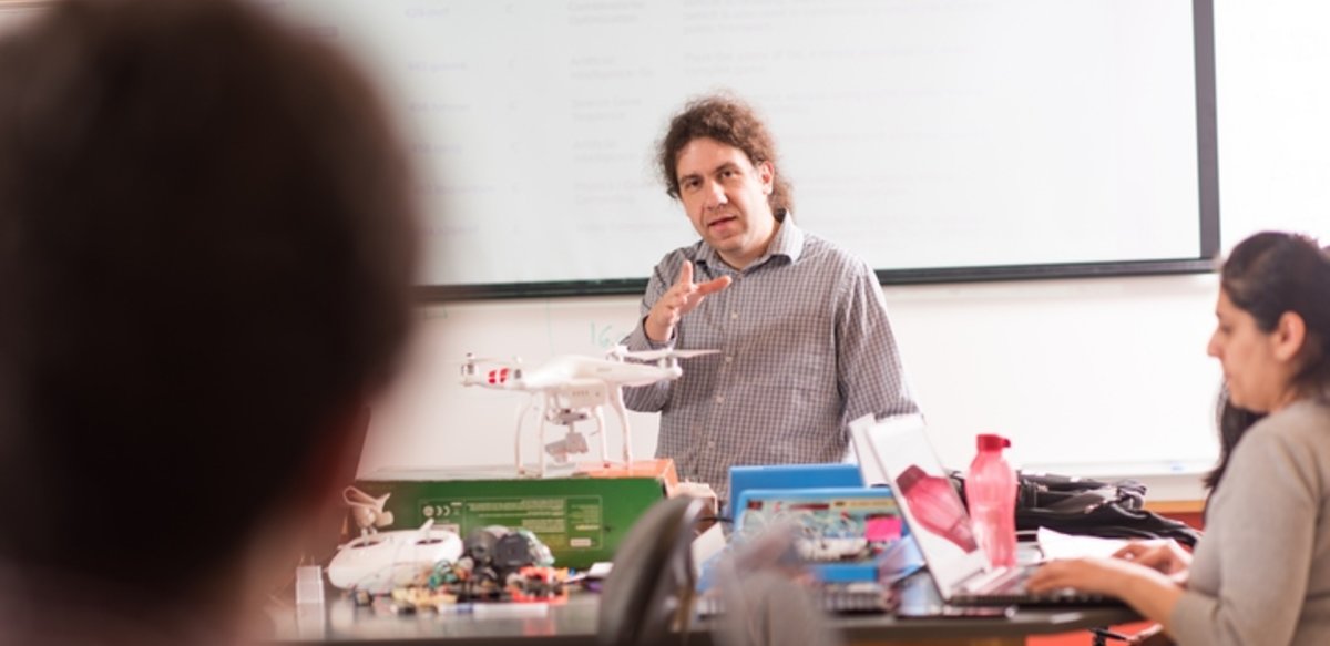 A professor speaking to a class in front of a projection screen and a desk with a drone and electronics on top of it. A professor speaking to a class in front of a projection screen and a desk with a drone and electronics on top of it.
