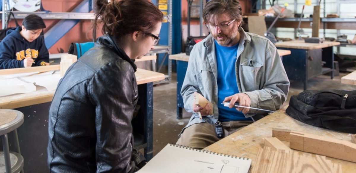 Woodworking classroom with the professor explaining something to one student and another student is working in the background. Woodworking classroom with the professor explaining something to one student and another student is working in the background.