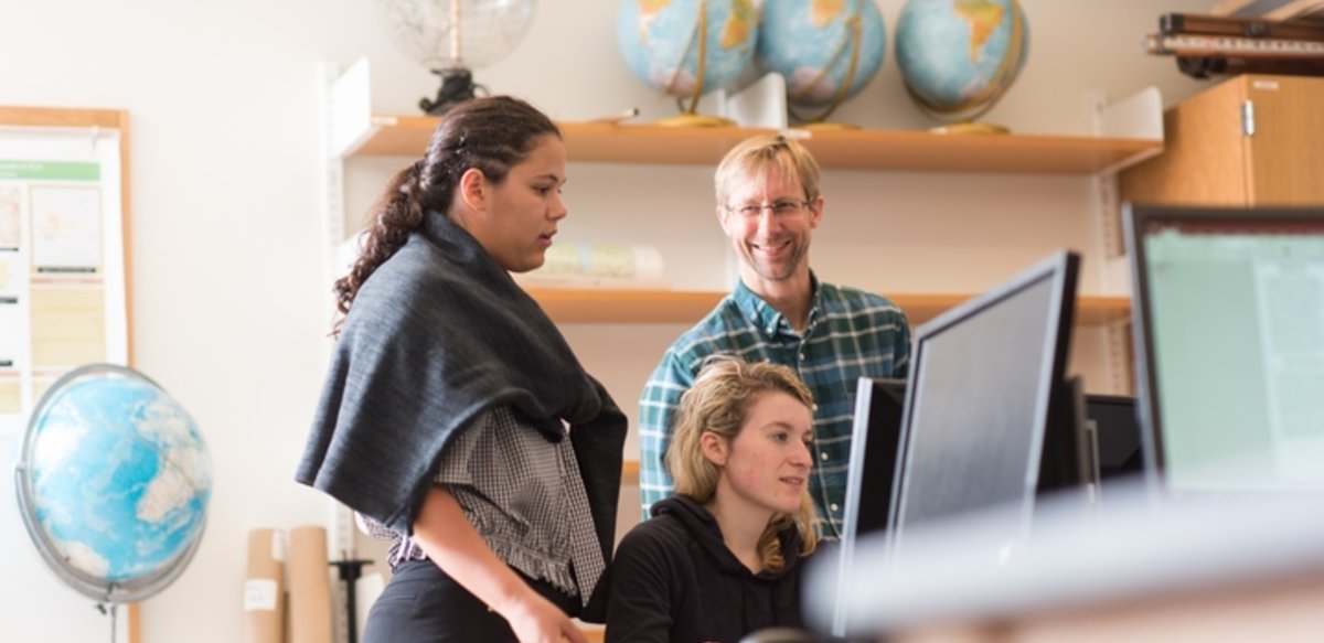 Professor Robert Hellstrom with 2 students at a computer in a classroom surrounded by globes.