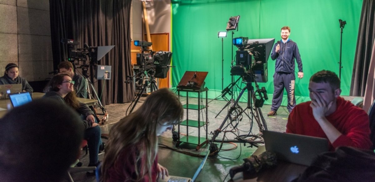 Foreground of students at tables working on laptops. Background of a student standing with a microphone in front of a green screen with tv cameras in front of him. Foreground of students at tables working on laptops. Background of a student standing with a microphone in front of a green screen with tv cameras in front of him.