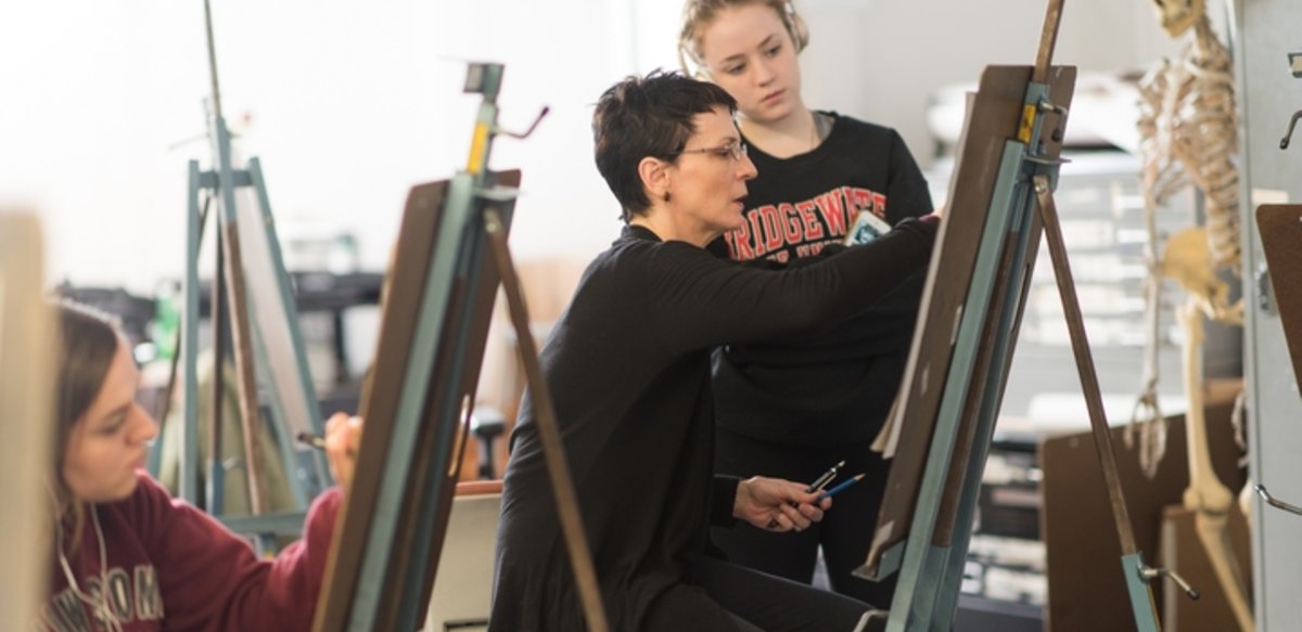 Drawing class with easels. One student sits drawing at an easel and a professor sits at the next easel teaching another student. Drawing class with easels. One student sits drawing at an easel and a professor sits at the next easel teaching another student.