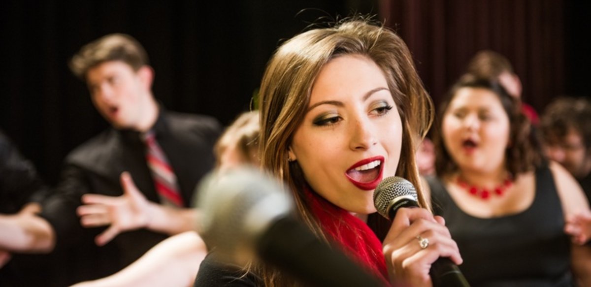 A vocal performance with a lead female student singer holding a microphone surrounded by other students singing and dancing. A vocal performance with a lead female student singer holding a microphone surrounded by other students singing and dancing.