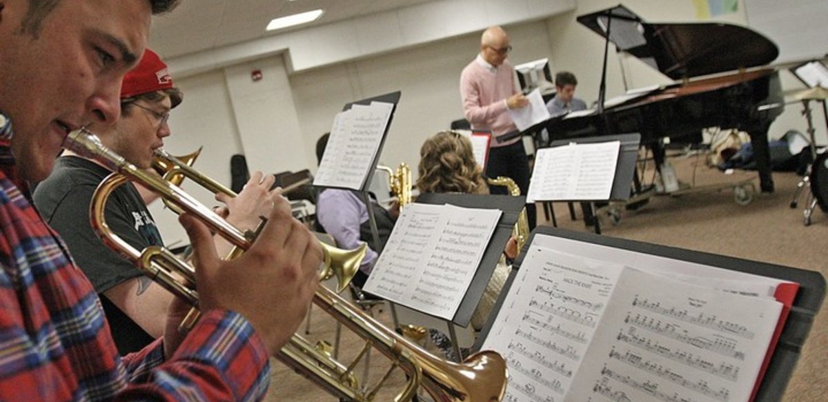 Jazz band class with students playing horns in the foreground, students playing saxophones, a professor, a piano and drum set in the background. Jazz band class with students playing horns in the foreground, students playing saxophones, a professor, a piano and drum set in the background.