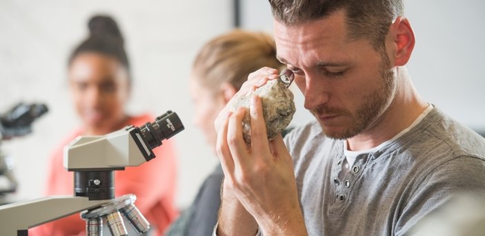 A student sitting in front of a microscope looking at a rock with a magnifier