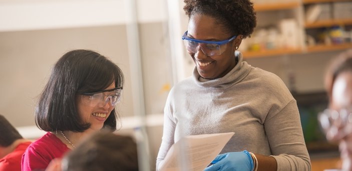 A student and professor, wearing rubber gloves and safety glasses, smiling and reviewing classwork.