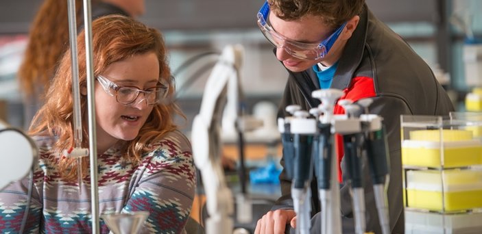 Two students working at a table in a chemistry lab wearing safety glasses with equipment in front of them.