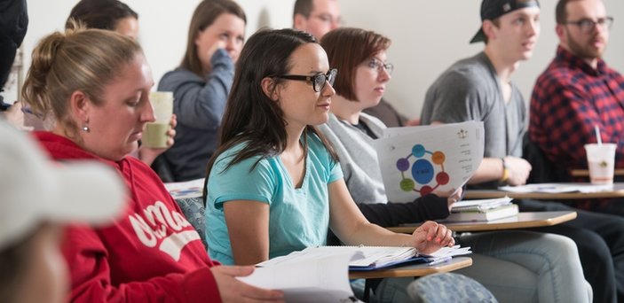 Students sitting in 2 rows of desks with notebooks out listening to a professor.