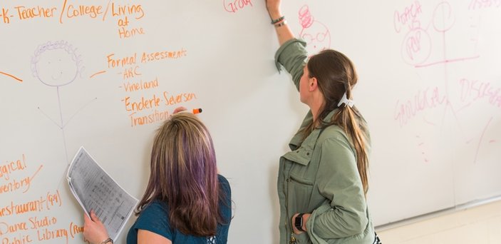 Two students writing with markers on a white board. Words on the board include "Formal Assessment" and "Coach vs. PCP."
