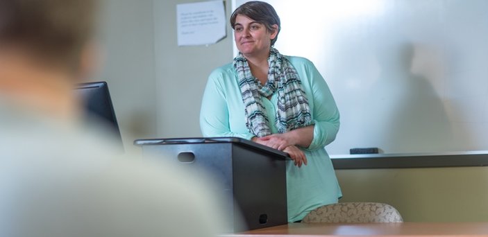 A professor teaching a class and smiling at a podium in front of a white board