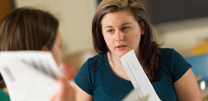 Two students at desks talking and holding coursework