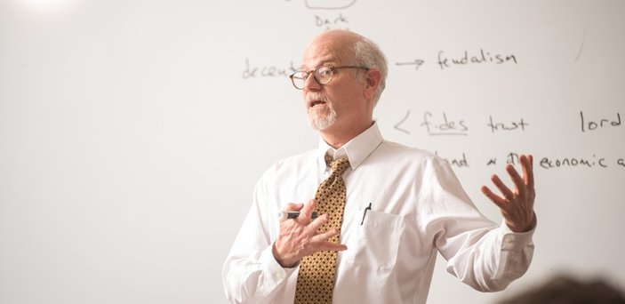 A professor teaching in front of a white board with words on it including Dark Ages, urbanism, feudalism and lord.