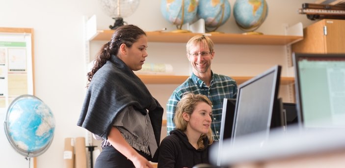 Professor Robert Hellstrom with 2 students at a computer in a classroom surrounded by globes.