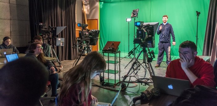 Foreground of students at tables working on laptops. Background of a student standing with a microphone in front of a green screen with tv cameras in front of him.