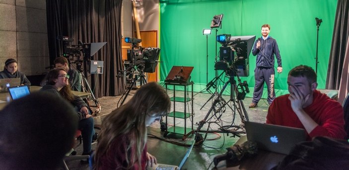 Foreground of students at tables working on laptops. Background of a student standing with a microphone in front of a green screen with tv cameras in front of him. 